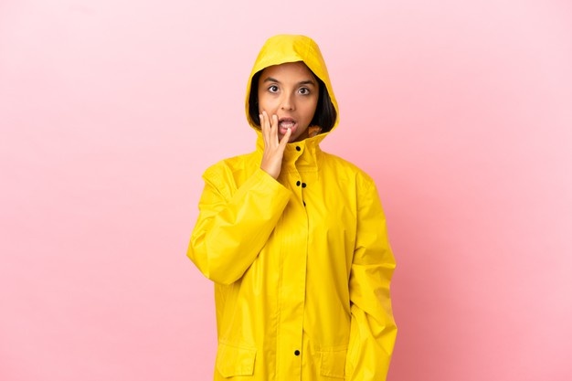 Молодая латинская женщина в непромокаемом пальто на изолированном фоне с удивленным и шокированным выражением лица